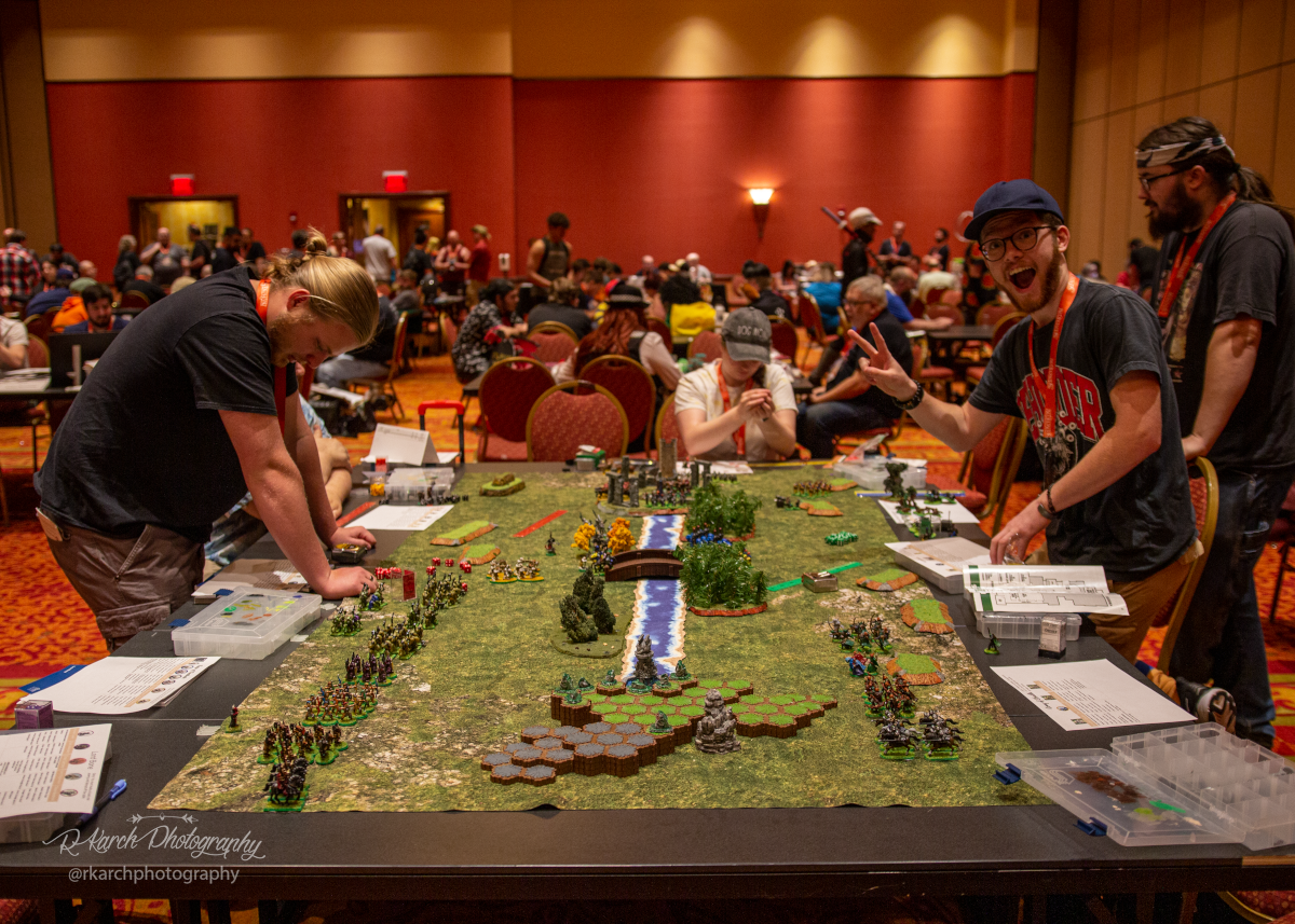 IMG_7150 Two players face off across the table over a miniature figures game. The man on right is looking at the camera giving a peace sign, the man on left is looking down at the table. There are many tabletop game players in the background.