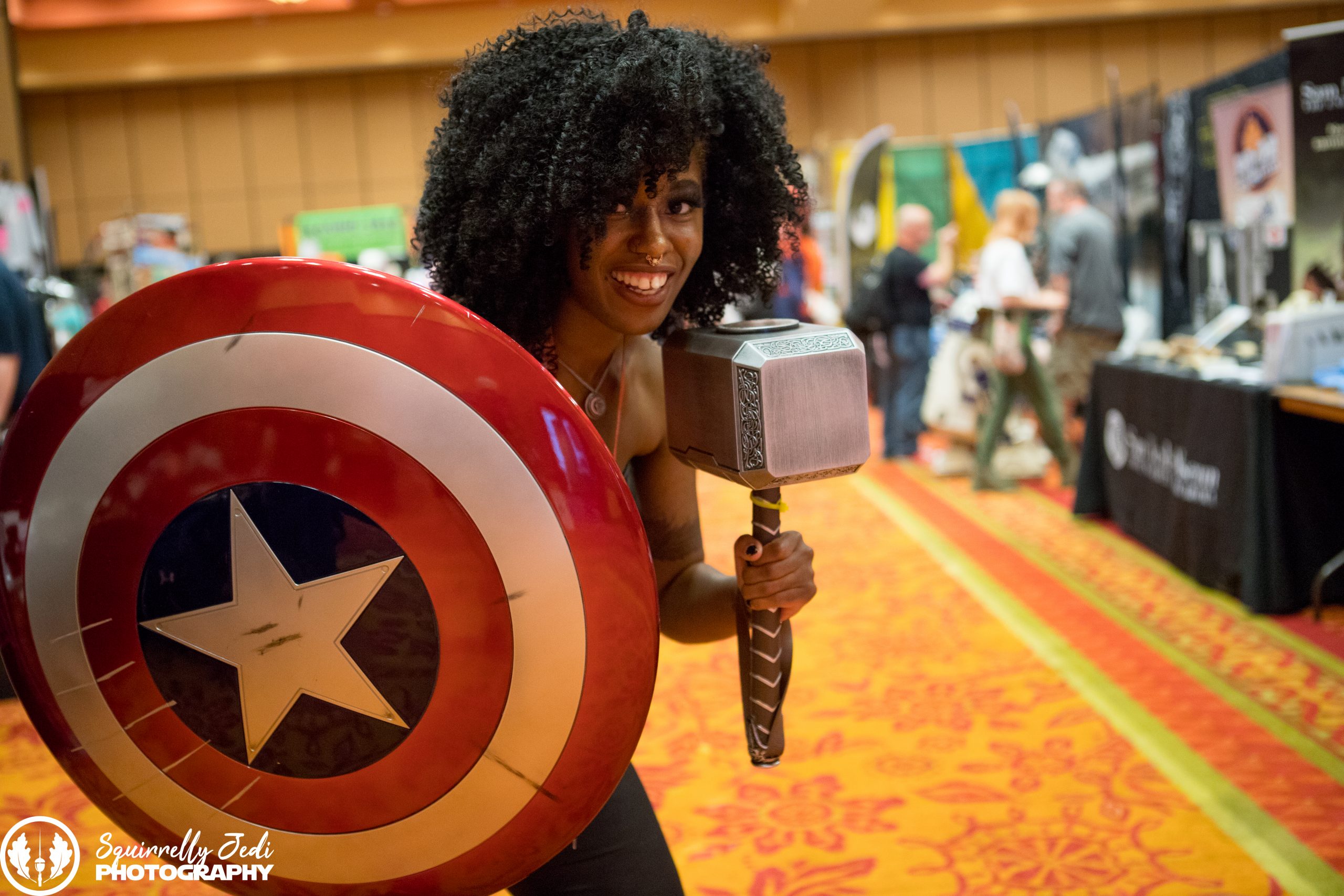 A woman smiles at the camera while holding a Captain America Shield and Thor's hammer.